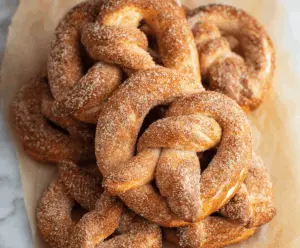 Golden-brown cinnamon sugar soft pretzels topped with a dusting of cinnamon sugar, fresh out of the oven, served on a rustic wooden surface.