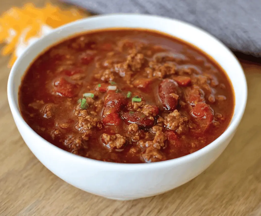 A delicious bowl of Copycat Wendy's Chili topped with shredded cheese and chopped onions, served in a white bowl on a rustic wooden table.