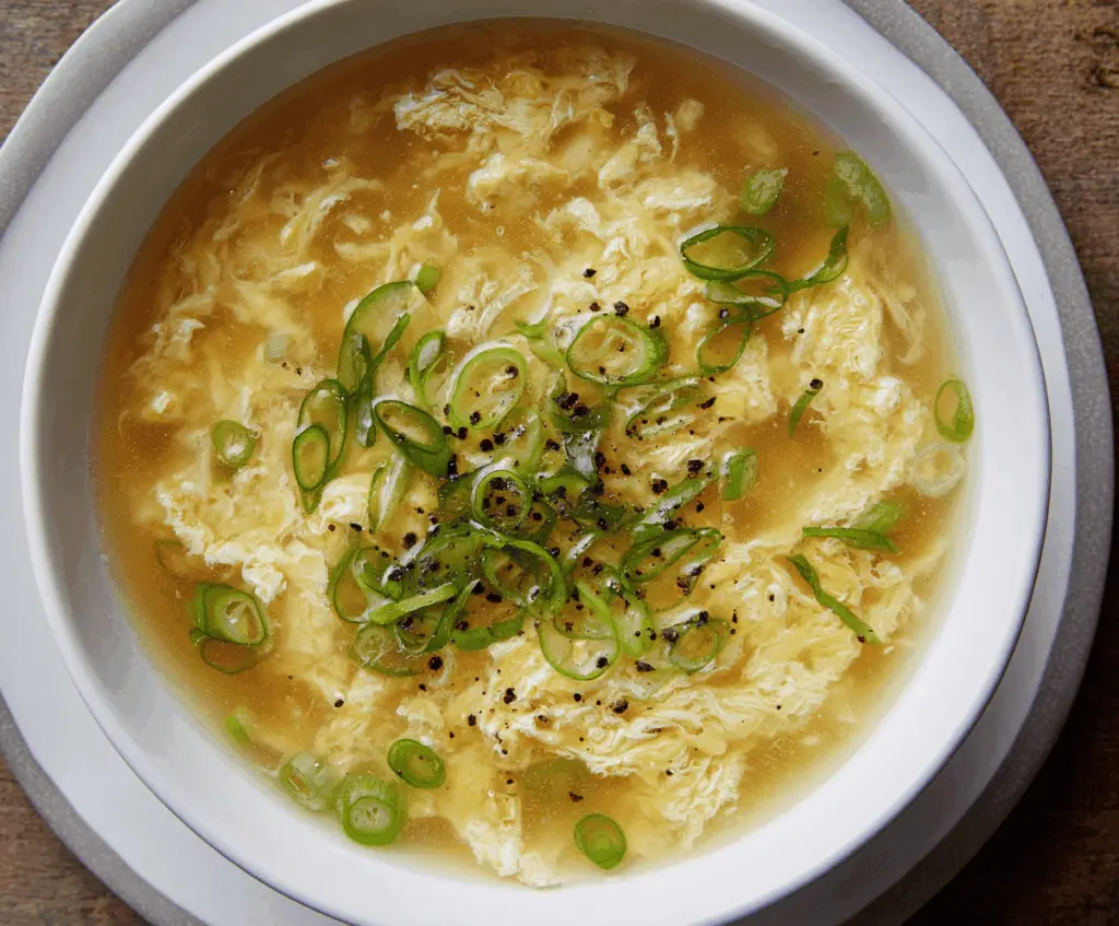 Creamy egg drop soup with tender egg ribbons, garnished with chopped green onions and served in a white bowl.